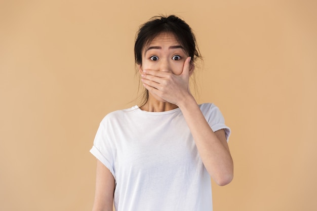 femme chinoise calme portant un t-shirt basique couvrant sa bouche avec les mains isolées sur un mur beige en studio