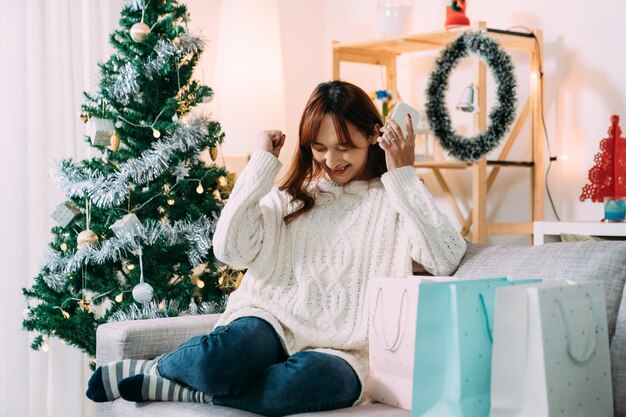 une femme chinoise crie de joie avec un poing levé pour gagner un prix tout en achetant des cadeaux de Noël en ligne sur son smartphone dans un salon confortable à la maison.