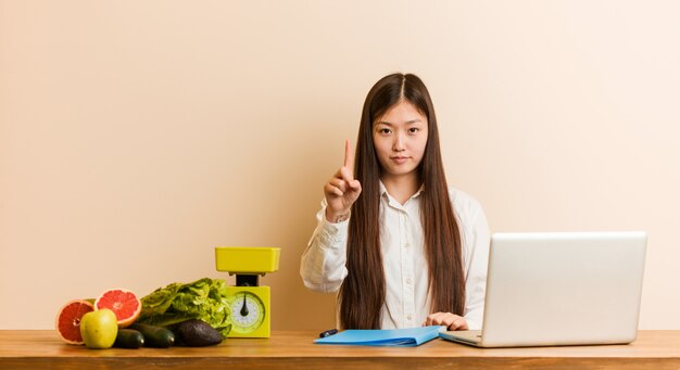 Femme chinoise jeune nutritionniste travaillant avec son ordinateur portable montrant le numéro un avec le doigt