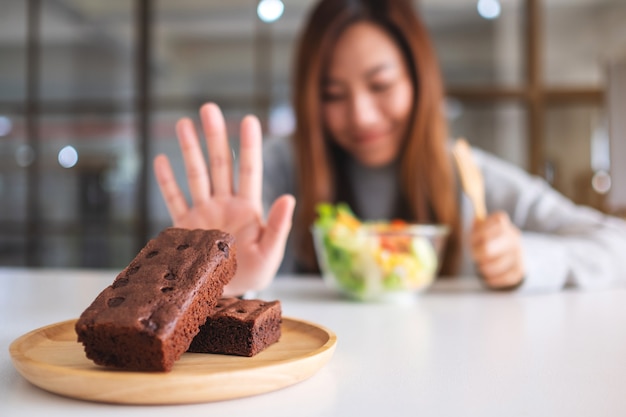 Une femme choisit de manger une salade de légumes et fait signe à la main de refuser un gâteau au brownie sur la table