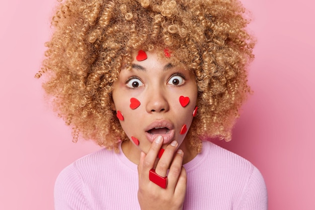 Une femme choquée aux cheveux bouclés retient son souffle a des cœurs sur le visage ne peut pas croire que ses yeux regardent les yeux sur écoute porte des vêtements décontractés isolés sur fond rose Réactions humaines et expressions du visage