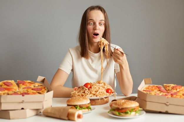 Femme choquée aux cheveux bruns portant un t-shirt blanc assis à table parmi des aliments malsains isolés sur fond gris posant avec une fourchette avec des pâtes se souvenant qu'elle garde son régime