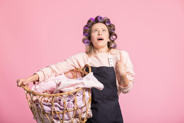 Une femme choquée lève les yeux vers l'espace vide destiné à la publicité pointe un doigt dans sa main tenant un panier en osier de linge publicitaire assouplissant sur fond rose