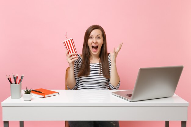 Une femme choquée, ravie, criant, écartant les mains tenant une tasse en plastique avec du cola ou du soda, travaille au bureau blanc avec un ordinateur portable isolé sur fond rose. Concept de carrière d'entreprise de réalisation. Espace de copie.