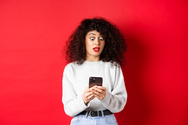 Femme choquée regarde l'écran du smartphone avec des yeux éclatés, lisant un message étrange, debout sur fond rouge.