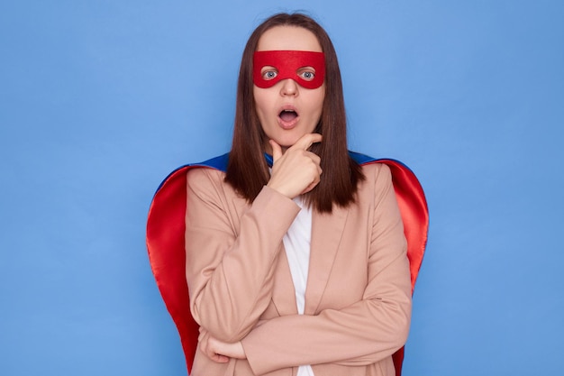 Une femme choquée et surprise portant un costume de super-héros se tient isolée sur un fond bleu tenant le menton regardant la caméra avec la bouche ouverte voit quelque chose d'étonné