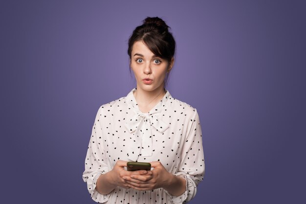 Une femme choquée tient un téléphone sur un mur de studio bleu en regardant la caméra dans une belle robe