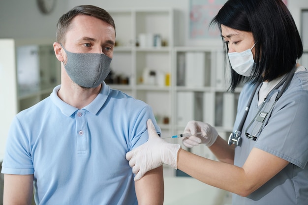 Femme clinicienne ou infirmière d'origine asiatique en uniforme et masque de protection vaccinant un jeune homme assis dans un cabinet médical de cliniques modernes