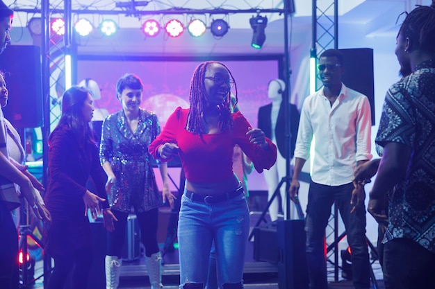 Femme clubber montrant des mouvements lors d'une bataille de danse improvisée pour divers amis dans une discothèque. Danseuse afro-américaine faisant la fête sur la piste de danse éclairée par des projecteurs de scène dans le club