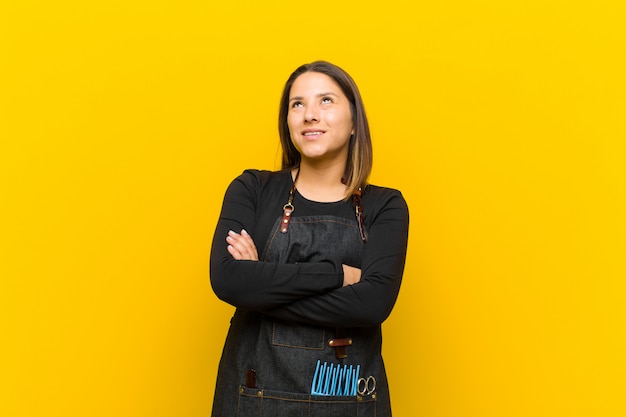 Femme coiffeur se sentir heureux, fier et plein d'espoir, se demandant ou pensant, levant les yeux pour copier l'espace avec les bras croisés contre le mur orange