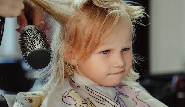 Une femme coiffeuse fait une jolie coiffure à la mode pour une jolie petite fille blonde