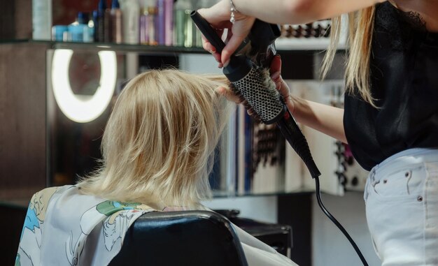 Une femme coiffeuse fait une jolie coiffure à la mode pour une jolie petite fille blonde