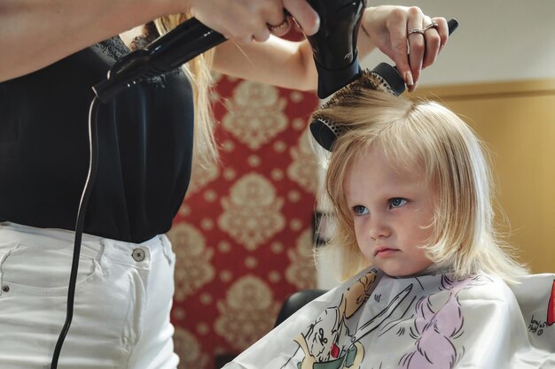 Photo une femme coiffeuse fait une jolie coiffure à la mode pour une jolie petite fille blonde