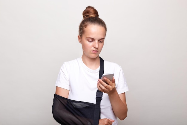 Femme avec une coiffure chignon portant un t-shirt décontracté posant isolé sur fond blanc a blessé le bras en écharpe utilise un téléphone portable pour faire défiler les réseaux sociaux trouve des conseils pour le traitement sur Internet