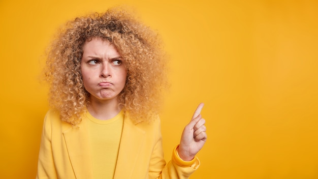 Une femme en colère aux cheveux bouclés regarde avec des points de jugement, le doigt à droite se tient debout, les joues bouleversées font une grimace irritée vêtue d'une tenue formelle isolée sur un mur jaune se plaint de quelque chose