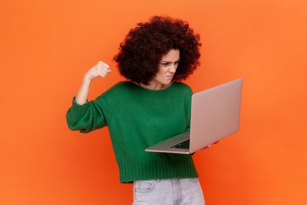 Femme en colère avec une coiffure afro portant un chandail de style décontracté vert travaillant sur un ordinateur portable a une erreur veut frapper son poing sur l'écran Prise de vue en studio intérieur isolée sur fond orange