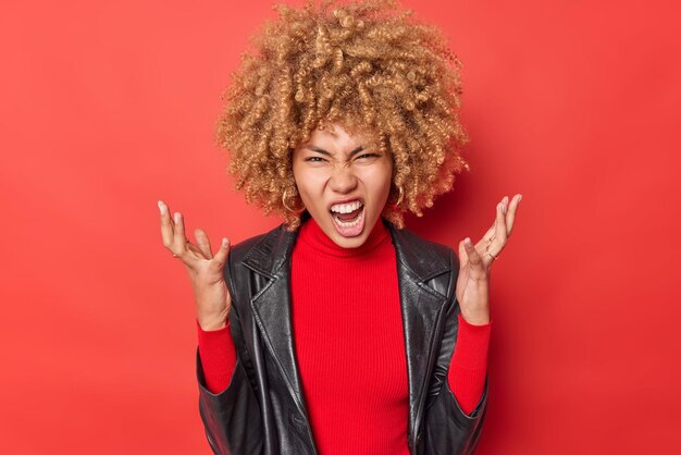 Une femme en colère outragée serre la main crie fort se sent ennuyée trie les relations se sent très ennuyée et énervée porte une veste en cuir noire isolée sur fond rouge Mauvaises émotions