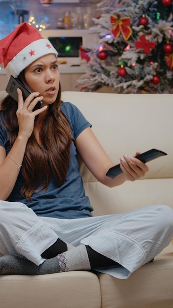 Femme en colère utilisant un smartphone pour travailler pendant les vacances de Noël. Personne portant un bonnet de noel, se sentant frustrée en parlant au téléphone et tenant la télécommande du téléviseur pour changer de chaîne