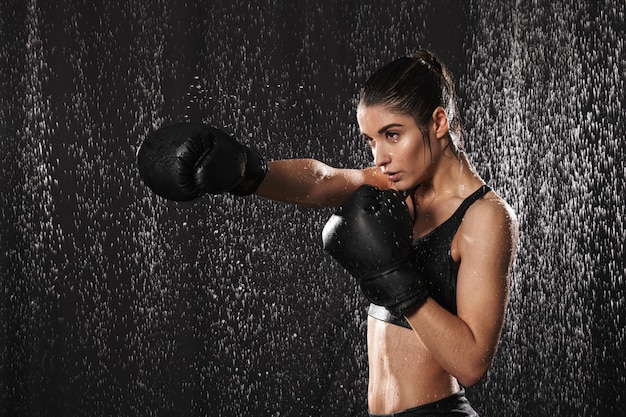 Femme combattant des années 20 dans les vêtements de sport et des gants de boxe noirs jetant des coups de poing sous des gouttes de pluie, isolé sur fond sombre