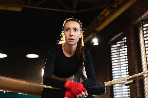 Femme combattante girl power Femme combattante avec des enveloppes de boxe rouges des bandages protecteurs debout sur un ring de boxe appuyé sur des cordes en attente de repos Forte fille puissante Force fit corps entraînement