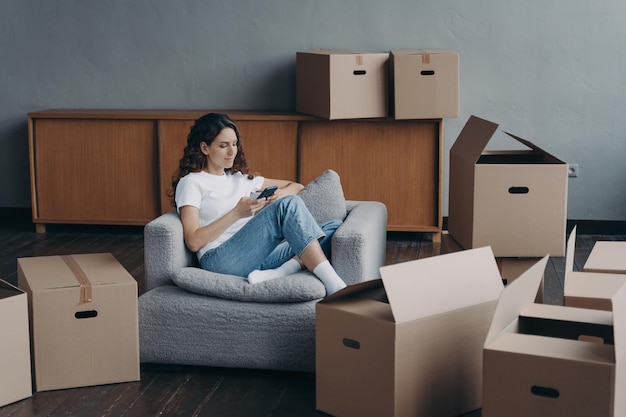 Une femme commande un service de déménagement à l'aide d'une application téléphonique le jour de la réinstallation, assise dans la chambre avec des boîtes en carton