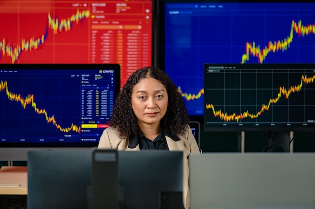 Une femme commerçante professionnelle à succès s'assoit souriante regarde la caméra entourée d'un écran d'ordinateur avec un stock de graphique et un rapport d'analyse de crypto-monnaie bitcoin dans la salle des marchés.