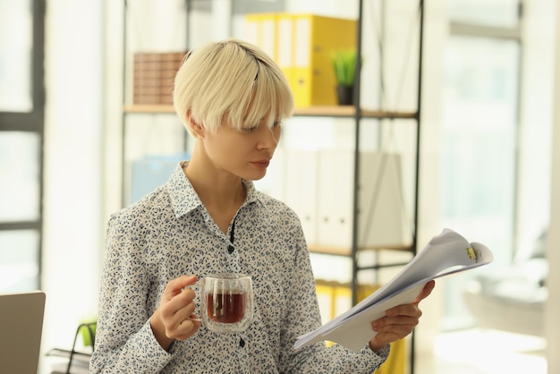 Femme concentrée aux cheveux blonds teints étudie les documents du projet tenant une tasse de thé jeune