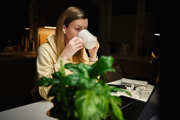 Une femme concentrée boit du café et regarde un écran d'ordinateur portable