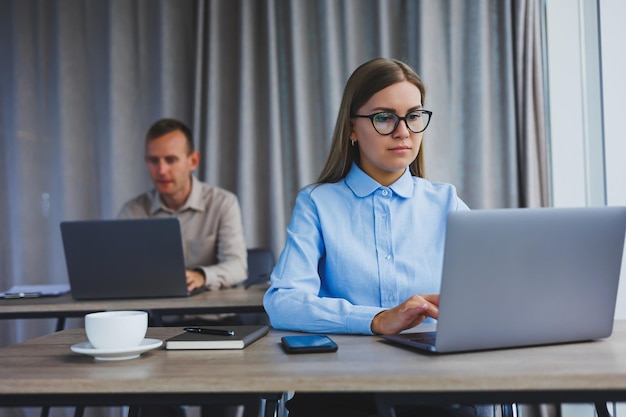 Femme concentrée dans des vêtements décontractés tapant sur un netbook tout en travaillant sur un nouveau projet tout en étant assise à un bureau dans un espace de travail moderne Femme d'affaires dans une veste et des lunettes Travail à distance