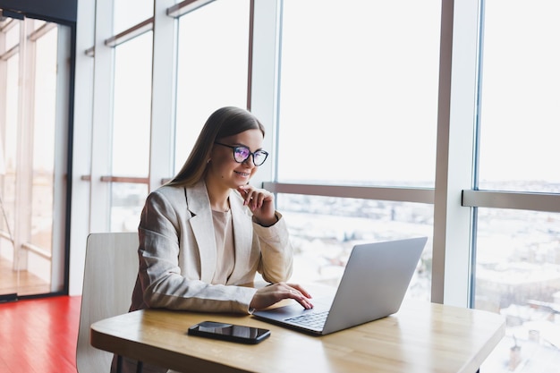 Femme concentrée en tenue décontractée tapant sur un netbook tout en travaillant sur un nouveau projet assis à un bureau dans un espace de travail moderne Femme d'affaires dans une veste et des lunettes Travail à distance