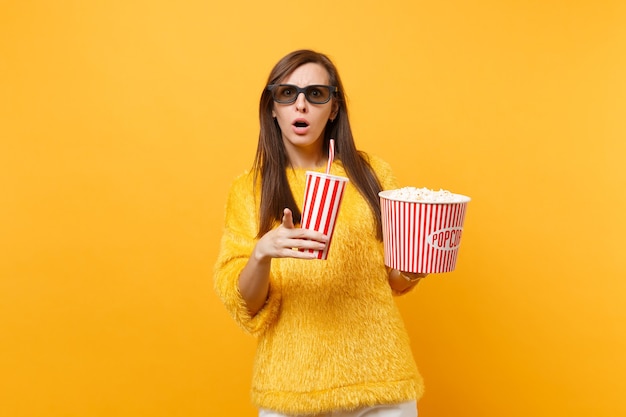 Femme concernée dans des lunettes imax 3d pointant l'index, regardant un film tenant un seau de pop-corn tasse de cola ou de soda isolé sur fond jaune. Les gens émotions sincères au cinéma, style de vie.