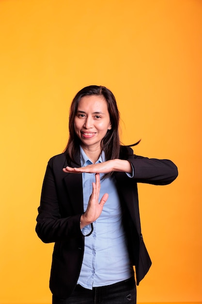 Femme confiante faisant le symbole de temporisation avec les mains, exprimant la mi-temps et la pause en studio sur fond jaune. Modèle asiatique souriant en costume formel demandant une pause, gesticulant signe de rejet.