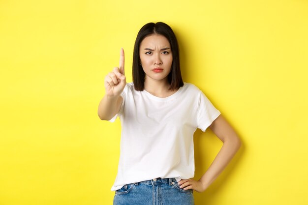 Femme confiante et sérieuse dit non, montrant le doigt étendu pour arrêter et interdire quelque chose de mal, fronçant les sourcils et regardant la caméra avec assurance, debout sur fond jaune.