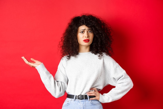 Femme confuse et sceptique aux cheveux bouclés, fronçant les sourcils et levant la main interrogée, ne peut pas comprendre quelque chose d'étrange, debout sur fond rouge