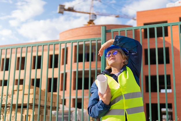 Femme constructeur sur chantier en gilet vert et casque bleu avec tablette Femme d'âge moyen avec des lunettes regardant la caméra Derrière c'est un bâtiment et une grue de construction