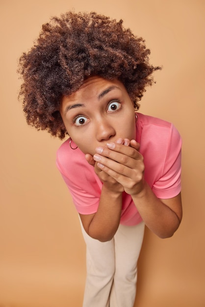 la femme contre la bouche essaie de se taire et de ne pas répandre de rumeurs porte un t-shirt rose décontracté isolé sur un beige regarde la caméra. Omg concept