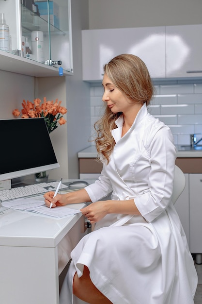 La femme cosmétologue vêtue d'une robe blanche est assise dans son bureau et remplit les documents