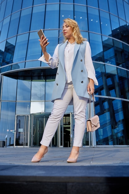 Femme en costume d'affaires se tient devant un immeuble de bureaux et regarde l'écran du smartphone