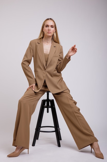 Femme en costume beige veste pantalon boutonné sur fond blanc Modèle assis sur une chaise