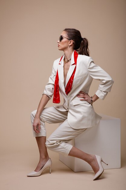 Femme en costume blanc veste pantalon chaussures accessoires rouges lunettes de soleil sur fond beige poudré