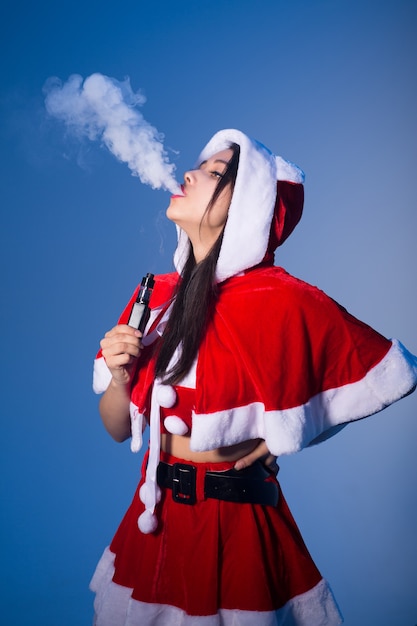 Femme en costume de père noël fumant une cigarette électronique et exhalant de la vapeur blanche sur fond bleu