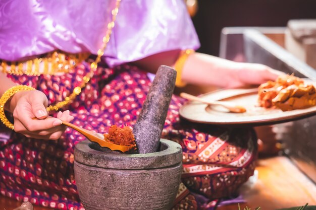 Une femme en costume traditionnel thaïlandais met une cuillère dans un plat pour récupérer la pâte de piment dans un mortier.