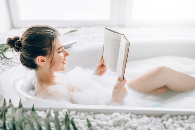 Femme couchée dans le bain avec de la mousse et lit le magazine