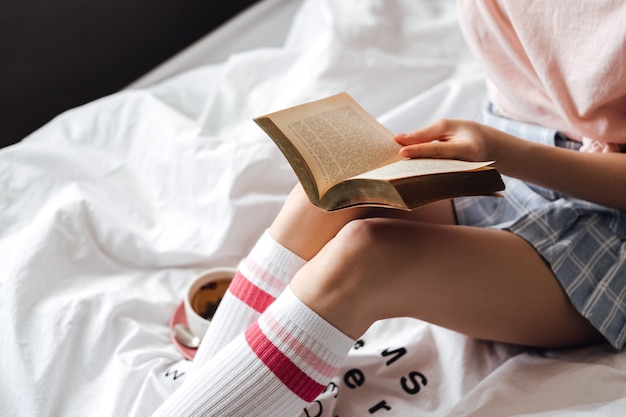 Femme couchée dans un lit blanc, boire du thé à la maison et lire un livre.