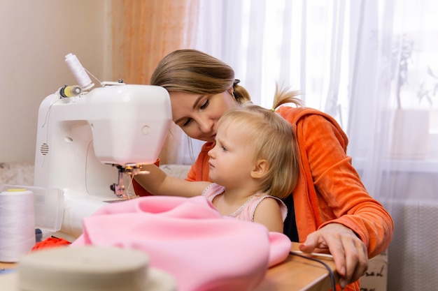 Une femme coud à la maison sur une machine à coudre électrique et s'engage avec sa petite fille