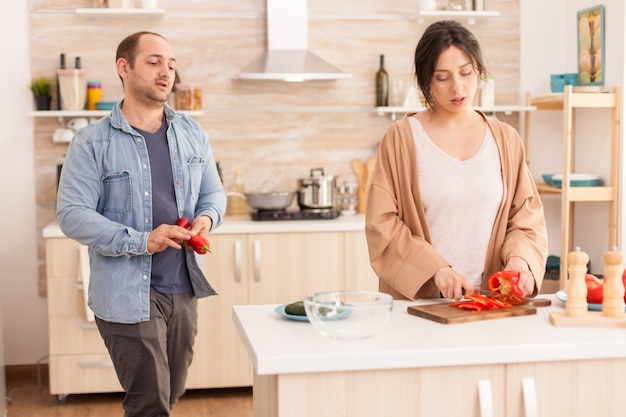 Femme coupant le poivron sur une planche à découper dans la cuisine. Mari tenant des légumes frais. Drôle de couple amoureux heureux à la maison, passer du temps ensemble, cuisiner sainement et souriant