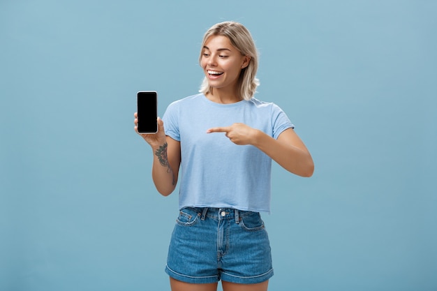 Femme avec coupe de cheveux blonde montrant l'écran du smartphone avec étonnement et plaisir en pointant l'appareil tout en faisant la promotion de tout nouveau gadget