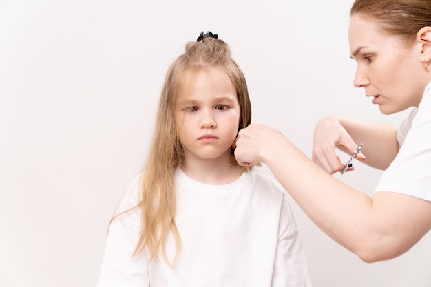 Une Femme Coupe Les Cheveux D'une Fille Drôle Sur Un Fond Blanc. Maman Coupe Les Cheveux De Sa Fille à La Maison. L'enfant A Peur Et Regrette Les Cheveux Coupés.