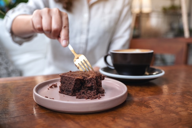 Une femme coupe gâteau brownie avec une fourchette avec une tasse de café sur une table en bois au café