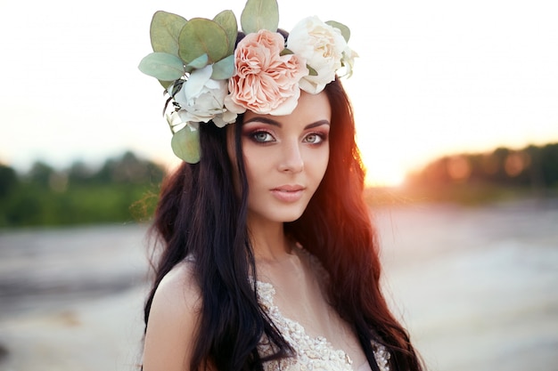 Femme avec une couronne de fleurs sur la tête et une robe d'été se promène dans les collines au coucher du soleil.